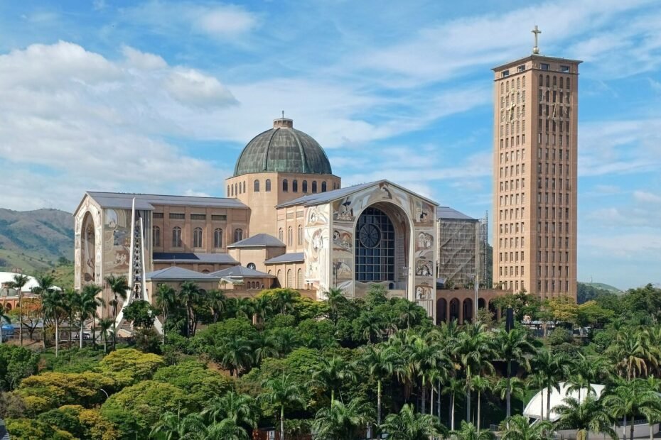 Basílica do Santuário Nacional de Aparecida, com cúpula e torre, cercada por palmeiras sob céu azul (SP)