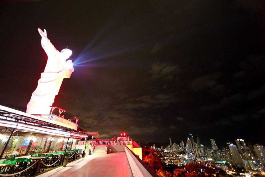 Estátua do Cristo Luz iluminada à noite, com os prédios acesos de Balneário Camboriú (SC) ao fundo