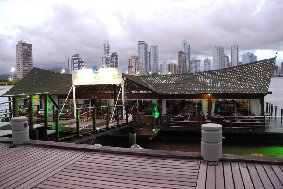 Restaurante flutuante iluminado ao entardecer, com os prédios da orla de Balneário Camboriú (SC) ao fundo