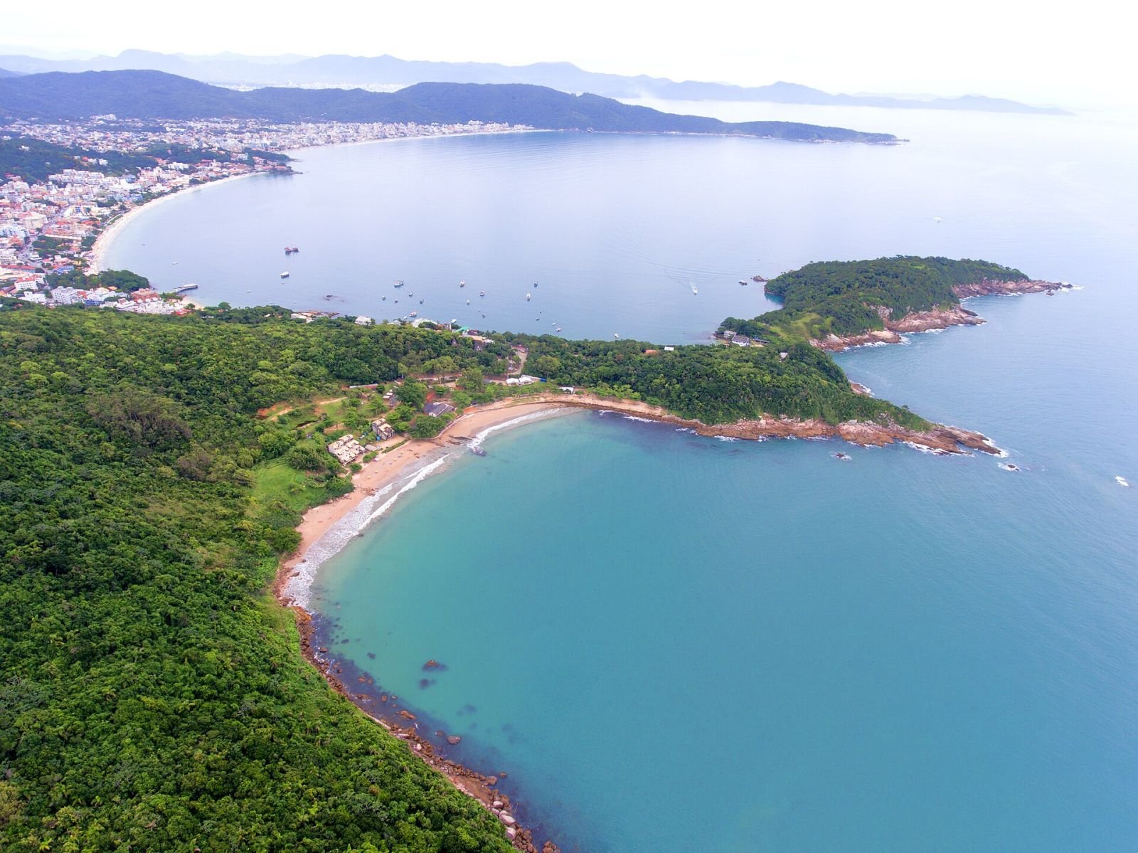 Vista aérea da Praia da Sepultura, enseada de águas azul-turquesa cercada de Mata Atlântica, em Bombinhas (SC)