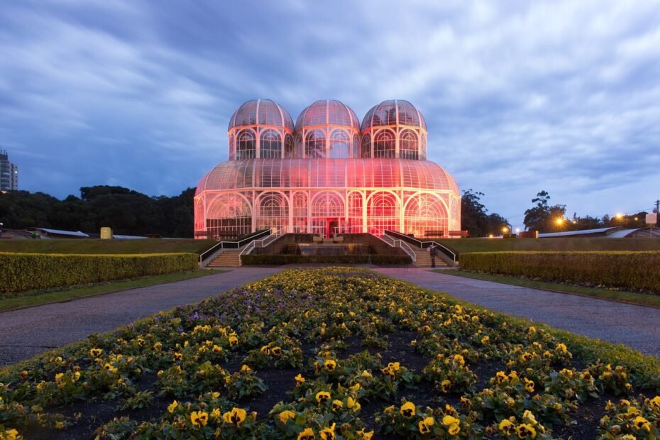Estufa de vidro do Jardim Botânico de Curitiba iluminada ao entardecer, com canteiro de flores amarelas em primeiro plano