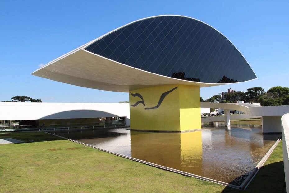 Prédio em forma de olho do Museu Oscar Niemeyer, apoiado em pilar amarelo sobre espelho d'água, em Curitiba