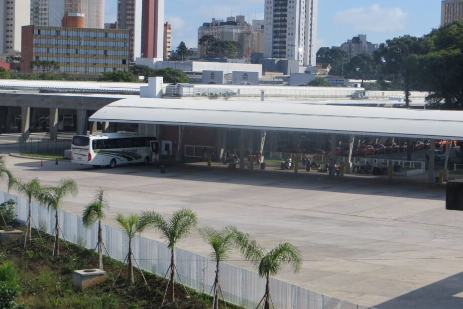 Plataformas cobertas da Rodoferroviária de Curitiba com ônibus de viagem e prédios do centro ao fundo