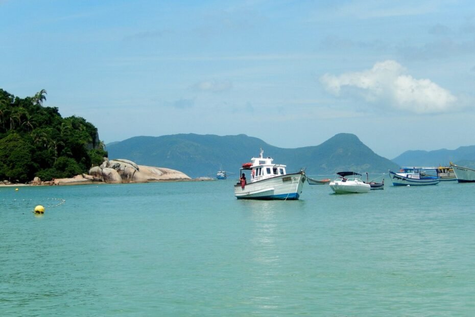 Barcos de passeio ancorados no mar verde-esmeralda da Ilha do Campeche, com morros ao fundo, em Florianópolis (SC)