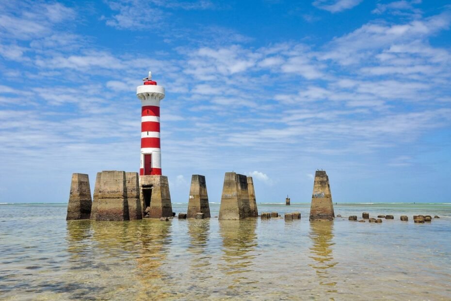 Farol listrado de vermelho e branco sobre pilares no mar raso da Praia de Ponta Verde, em Maceió (AL)