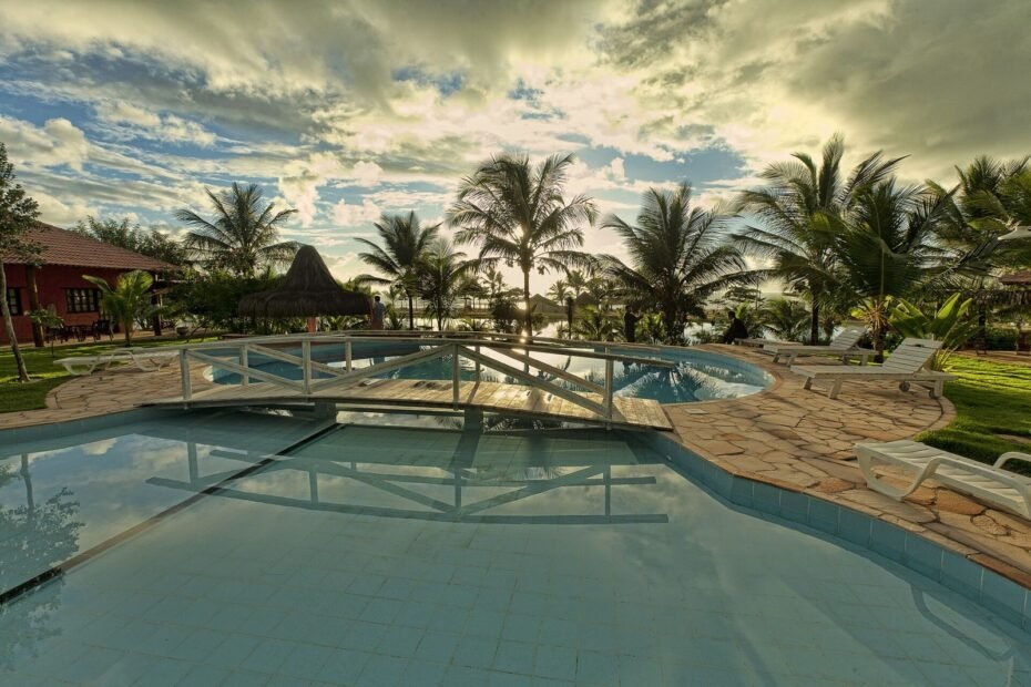 Piscina de resort com ponte de madeira, espreguiçadeiras e coqueiros ao amanhecer, na Costa das Baleias (BA)