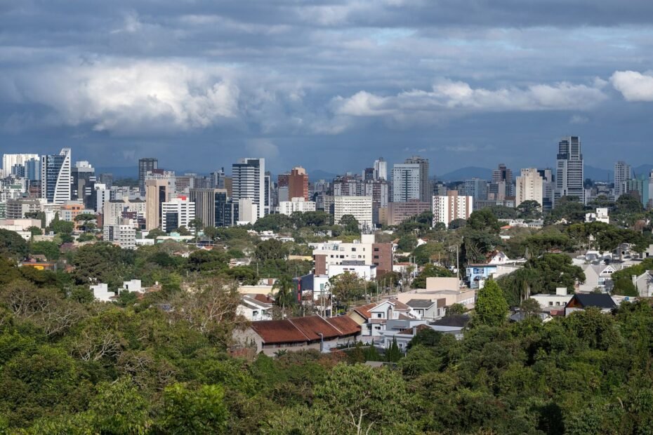 Vista panorâmica de Curitiba com prédios altos, casas e muito verde sob céu com nuvens